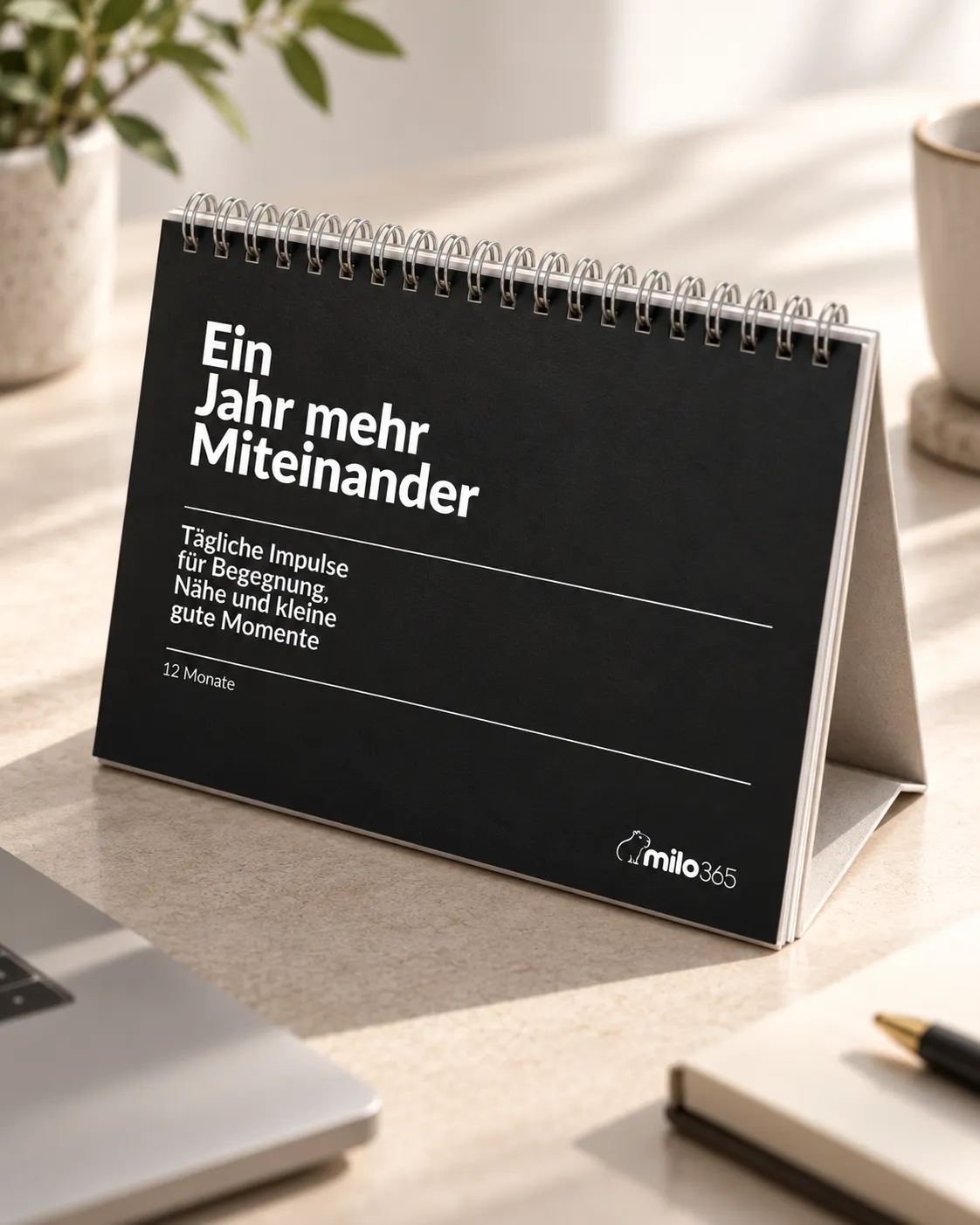Der Kalender mit schwarzem Cover auf einem sonnigen Schreibtisch, daneben Laptop, Pflanze und Tasse